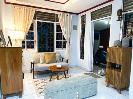 A room in the same house after staging: a sofa with cushions, a round coffee table on a patterned rug, an upholstered bench, wooden cabinets, a lit lamp and curtains.